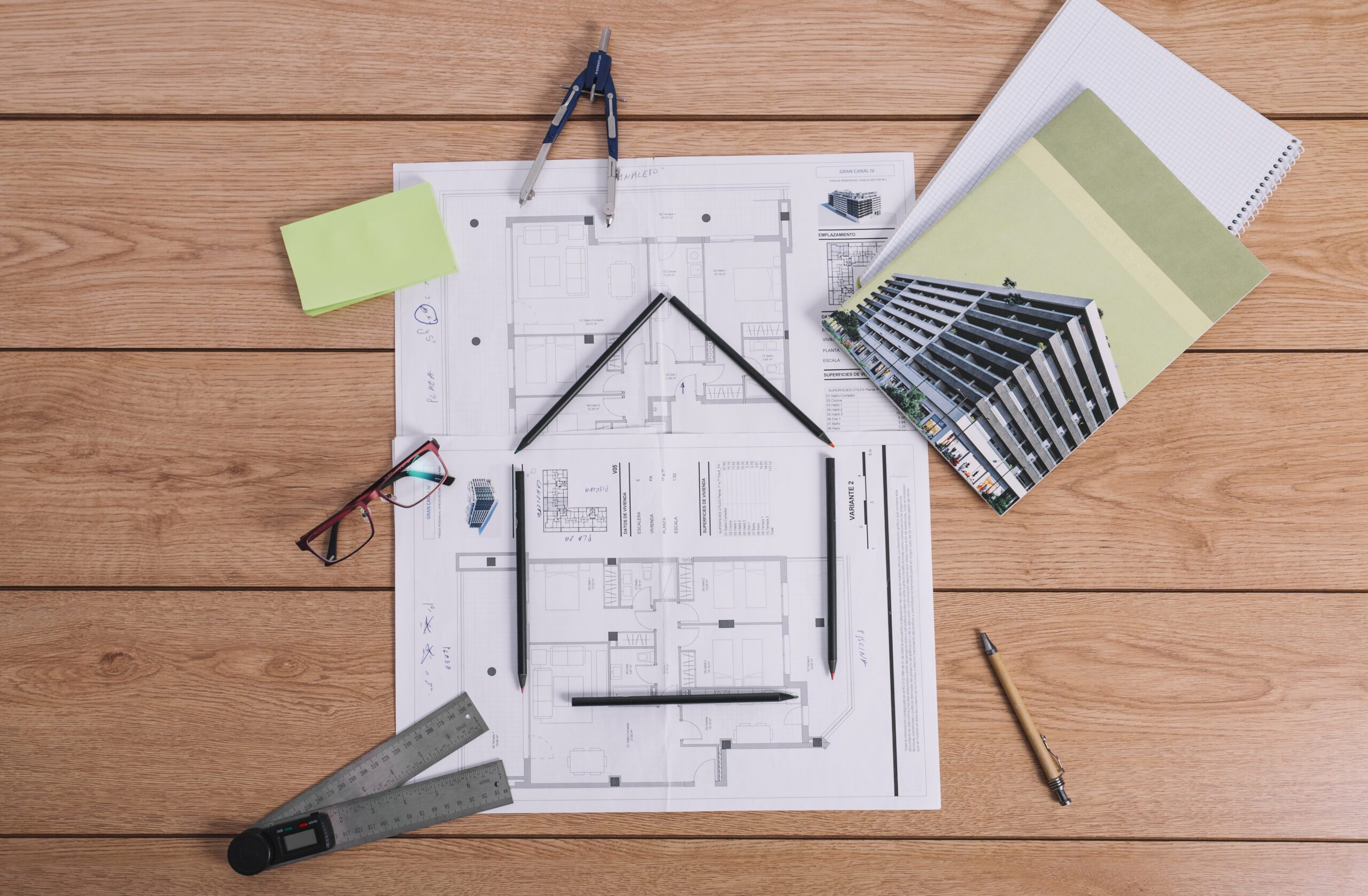 Vue de dessus d’un bureau. On y voit des plans de bâtiments posés sur une table en bois, entourés d’outils de dessin technique tels qu’un compas, des crayons, une équerre, une règle pliante, et des lunettes. Un carnet avec une photo d’un immeuble moderne est posé à droite, accompagné d’un cahier quadrillé. Un post-it vert et un stylo complètent la scène. L’ensemble évoque un environnement de travail en architecture ou en ingénierie du bâtiment.