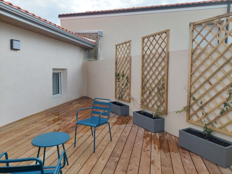 Une terrasse en bois avec deux chaises et une petite table bleues. Trois jardinières rectangulaires grises sont alignées contre un mur clair, chacune avec un treillis en bois où grimpent de jeunes plantes.