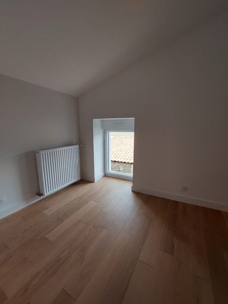 Une pièce lumineuse avec parquet en bois clair, un radiateur blanc fixé au mur et une petite fenêtre donnant sur des toits en tuiles rouges.