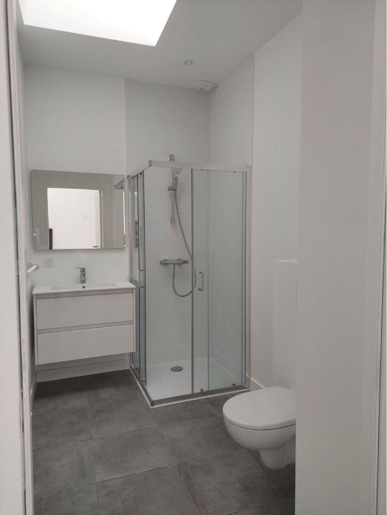 Salle de bain moderne avec douche vitrée, meuble vasque design suspendu, toilettes murales et carrelage gris contemporain.