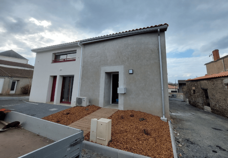 Une maison à deux niveaux, avec une façade clair et un toit en tuiles. L’entrée est accessible par un petit chemin en bois entouré de paillage et de jeunes plantations.